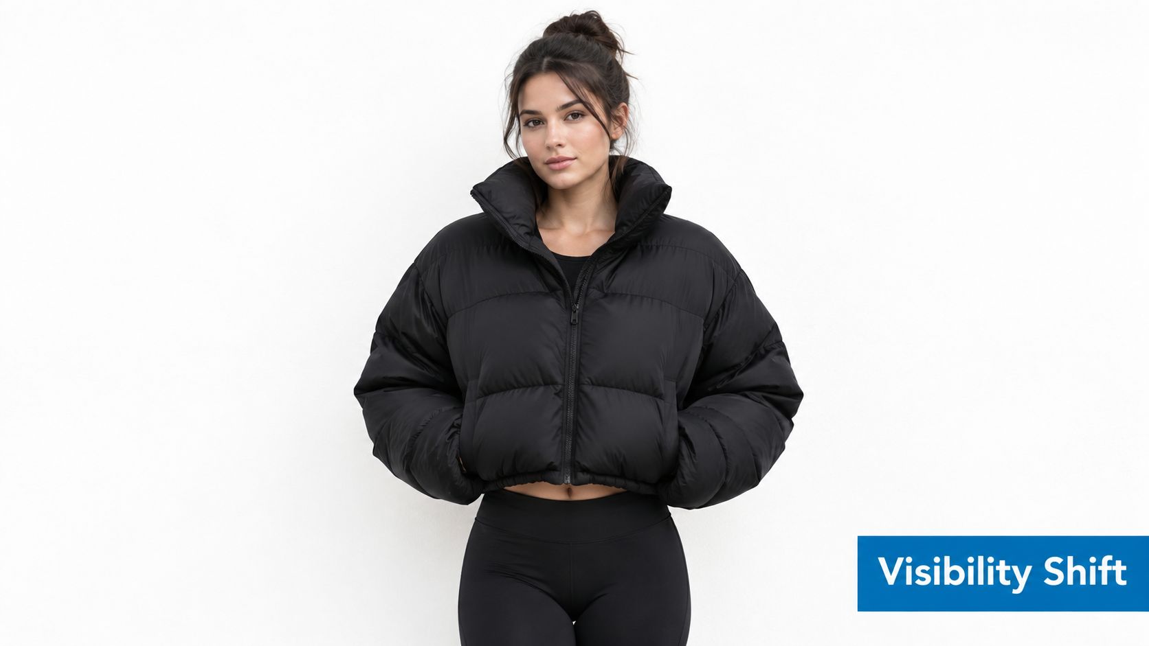 A young woman wearing a black cropped puffer jacket stands against a plain white wall background.